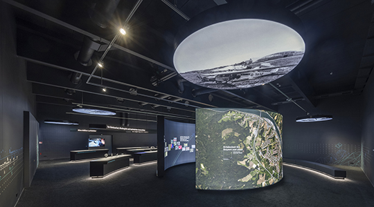Der Blick in die Dauerausstellung des Bayerischen Landesluftbildzentrums zeigt eine gebogene Wand auf der Luftbilder dargestellt sind. Über der Wand ist ein Spiegel angebracht. Der Raum ist dunkel gestrichen und es sind im Hintergrund weitere Exponate zu sehen.